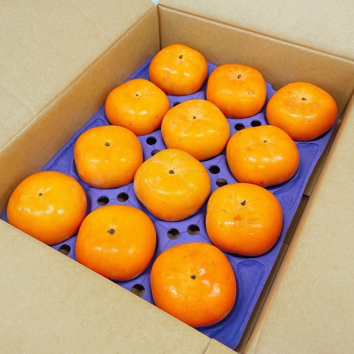 
                  
                    Twelve Akiou Japanese persimmons arranged in a purple protective tray inside an open cardboard box
                  
                